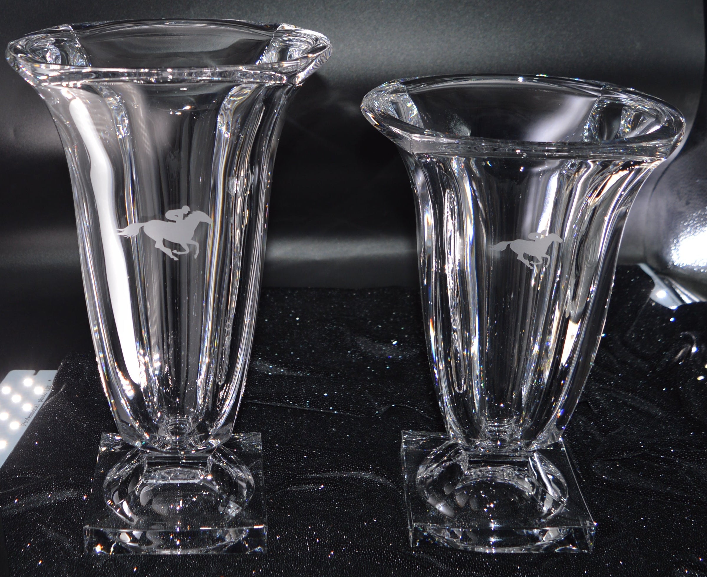 Two clear glass vases on a reflective black surface with a metallic object in the background.