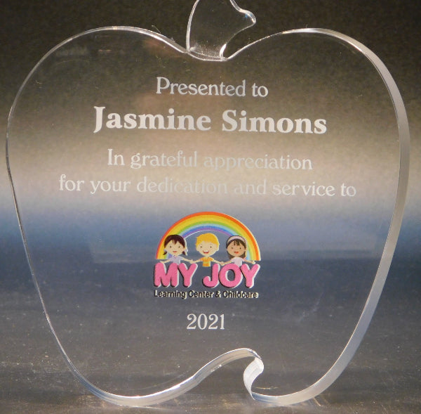 Clear acrylic apple-shaped award with engraved text and logo on a reflective surface