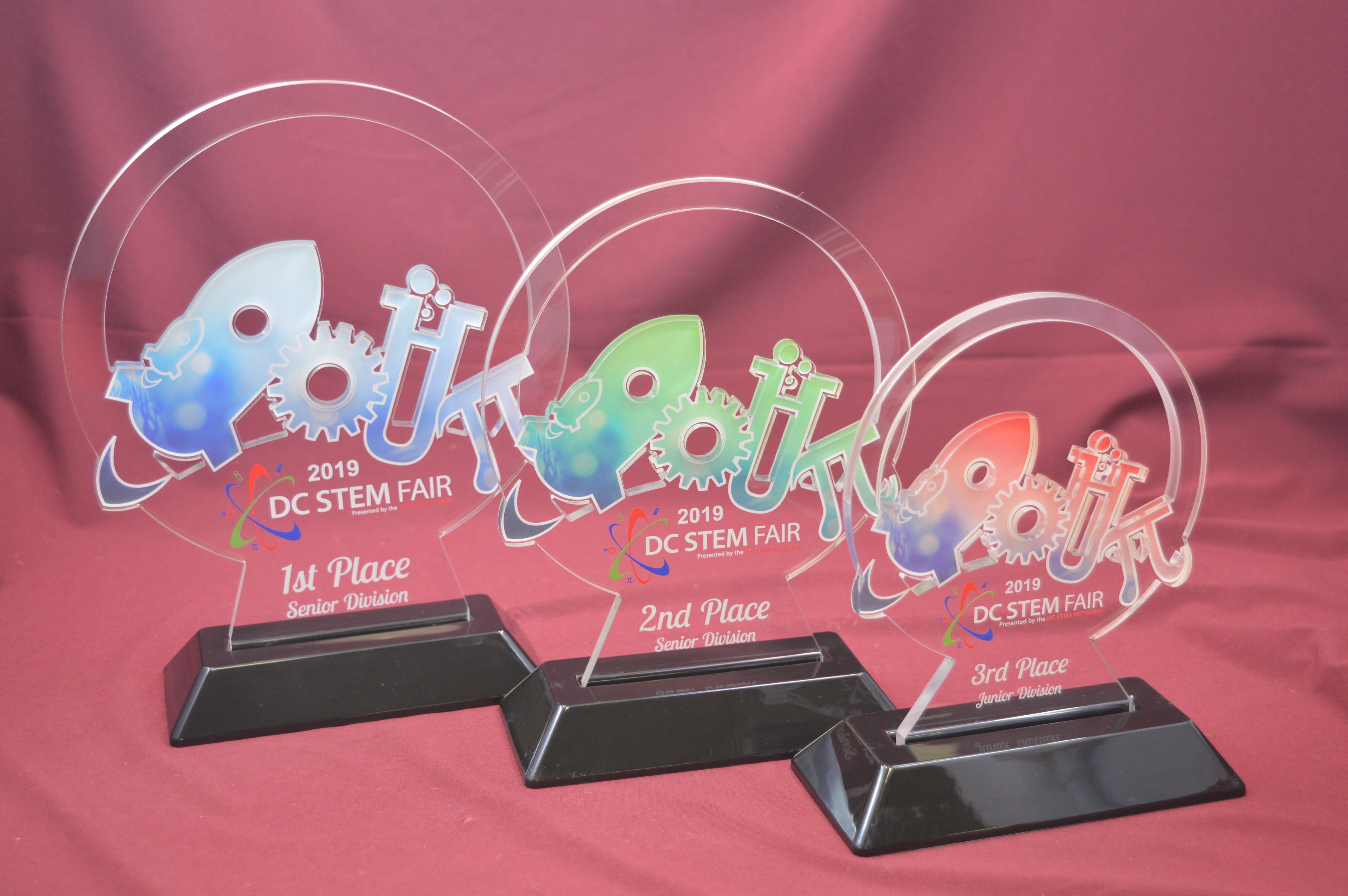 Three colorful acrylic awards for a DC STEM Fair on a red background